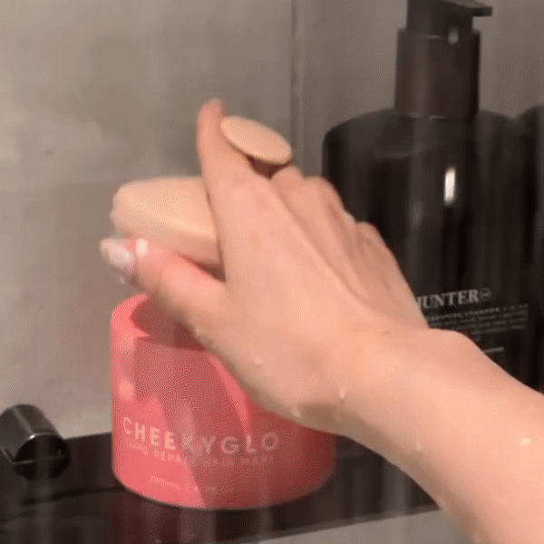A hand reaches to place a pink, round exfoliating pad back onto a jar labeled "CheekyGlo" in a bathroom setting. Adjacent to the jar is a black pump bottle labeled "Hair & Scalp Set" from CheekyGlo, suitable for all hair types. Water droplets are visible, indicating a recent shower or bath.