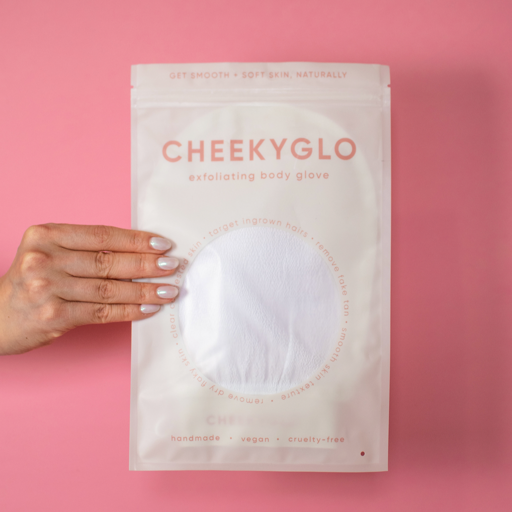 A hand with pale pink nails displays the CheekyGlo Sensitive Exfoliating Glove against a pink background. The packaging is white, adorned with red text that says, "Get smooth + soft skin, naturally," and highlights features such as "handmade," "vegan," and "cruelty-free." It&