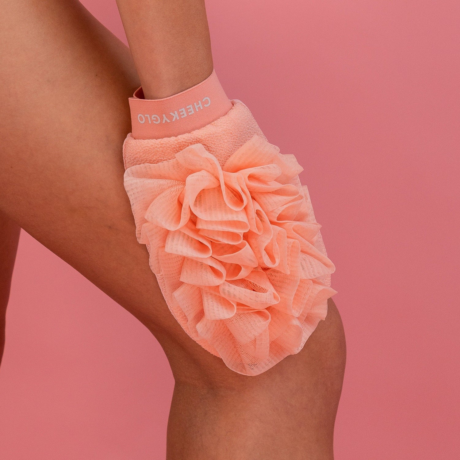 A person using a soft, peach-colored CheekyGlo Exfoliating Loofah Glove with a ruffled texture on their leg. The glove, branded "CheekyGlo" on the wrist strap, promises to help with keratosis pilaris and leave the skin smooth. The background is a solid pink color.