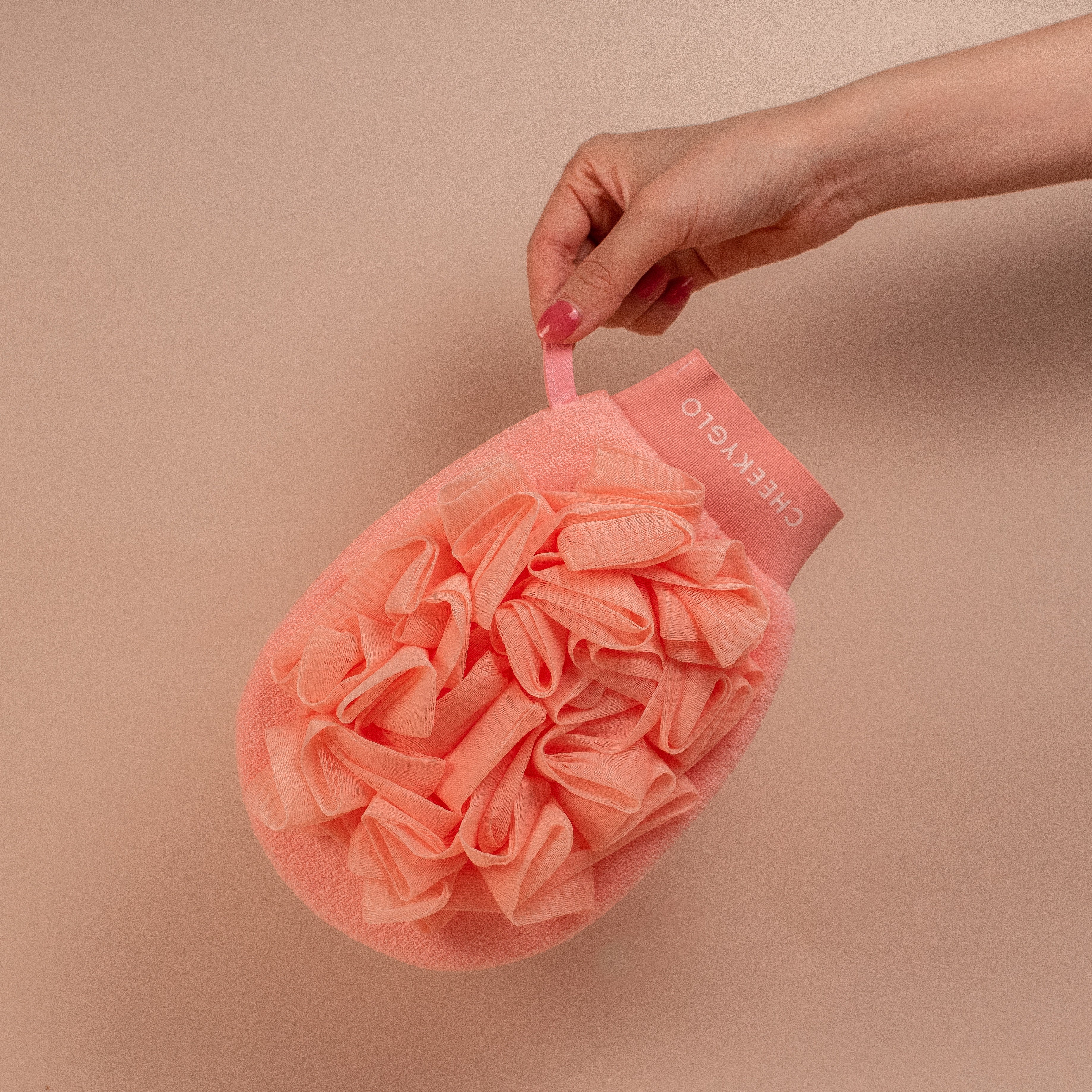 A hand holding a pink CheekyGlo Exfoliating Loofah Glove with a strap labeled "CHEEKYGLO." The glove features a textured, ruffled surface and an oval shape. Ideal for tackling keratosis pilaris, it promises skin-smoothing results. The background is a plain beige color.