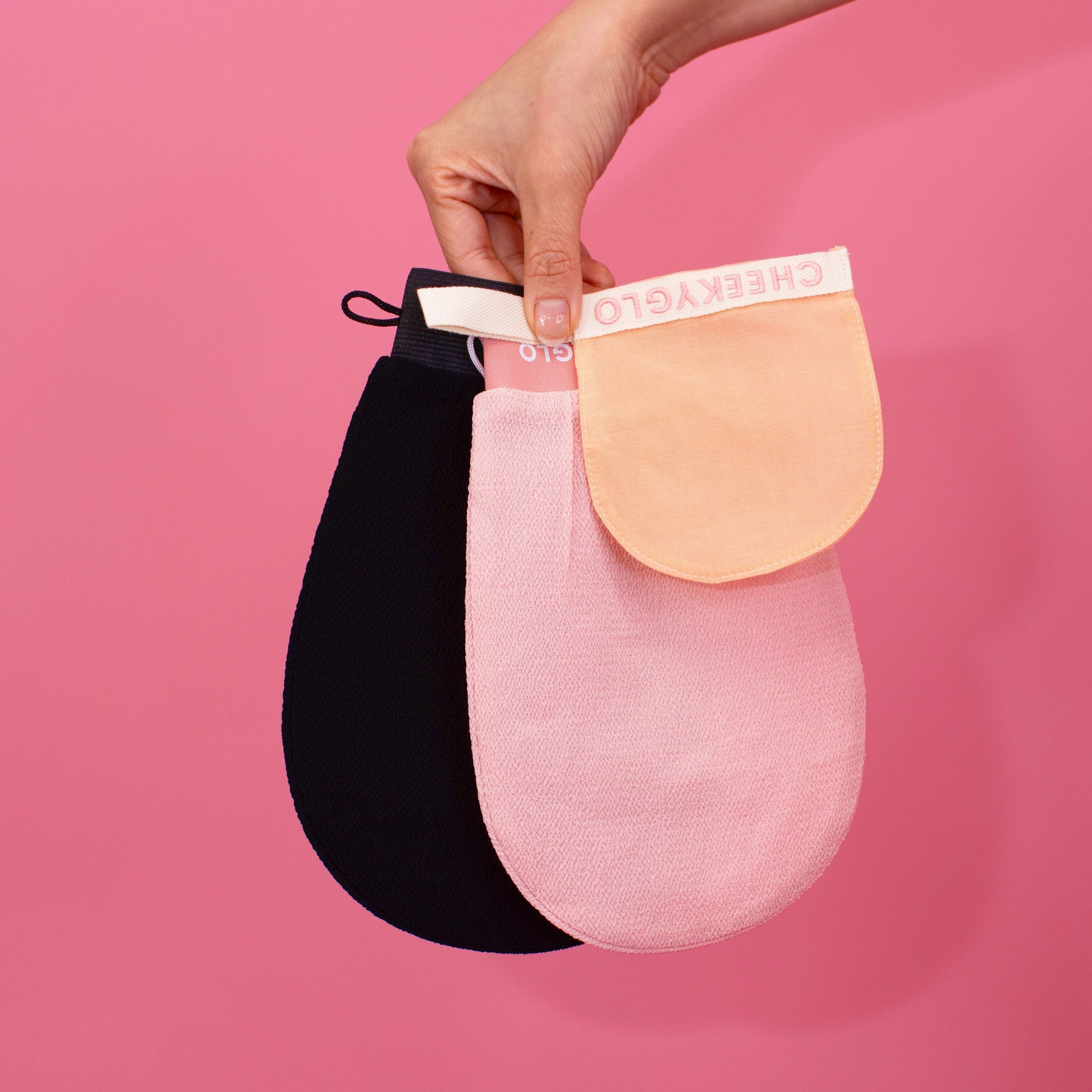 A hand holding three exfoliating gloves from the CheekyGlo Exfoliating Trio against a pink background: one black, one light pink, and one beige. Each oval-shaped body glove features a strap labeled "CHEEKY GLO".