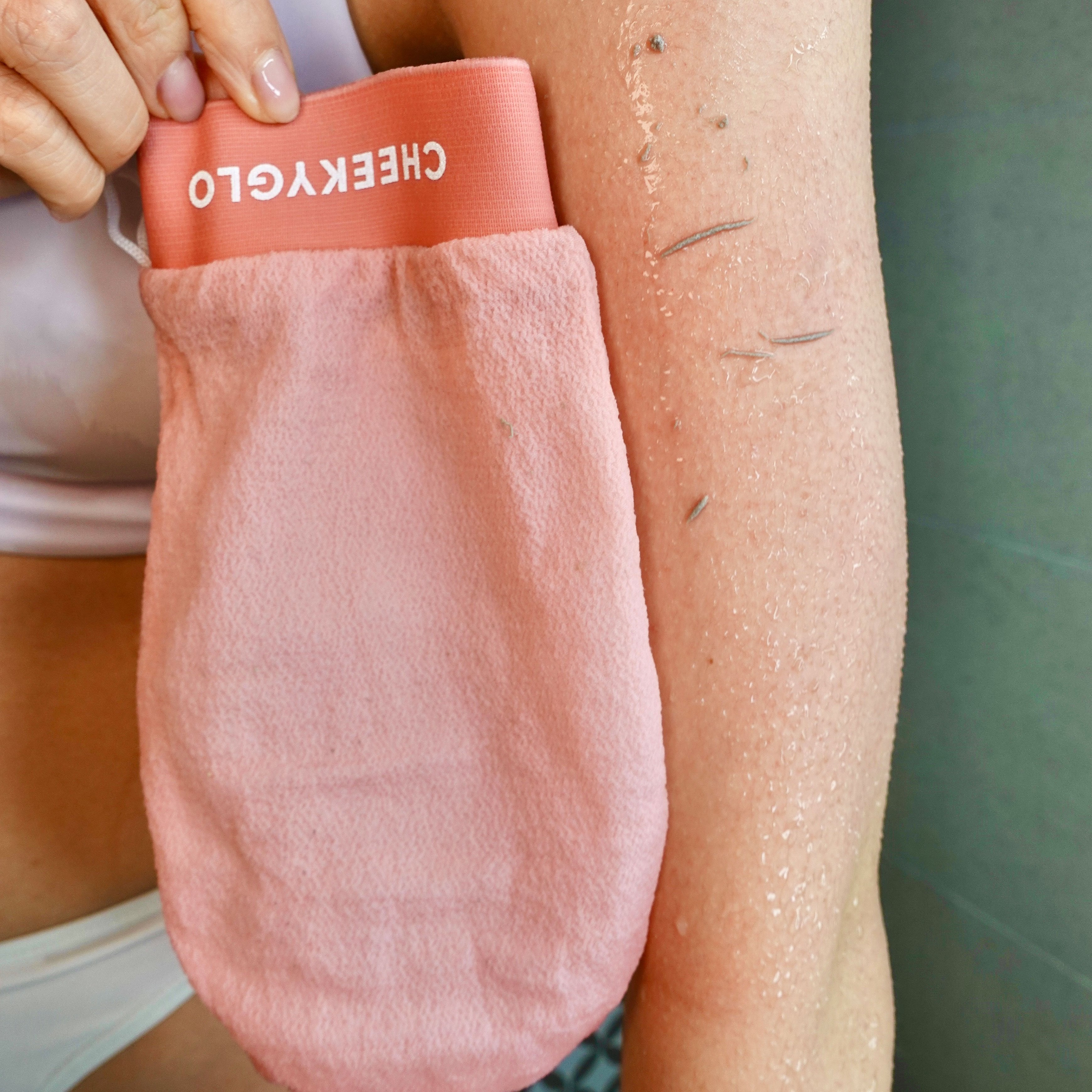 A person in a white top is using a pink exfoliating glove from the "Exfoliating Trio" by CheekyGlo to remove dead skin cells from their arm. The body glove shows visible particles of exfoliated skin, revealing its effectiveness.