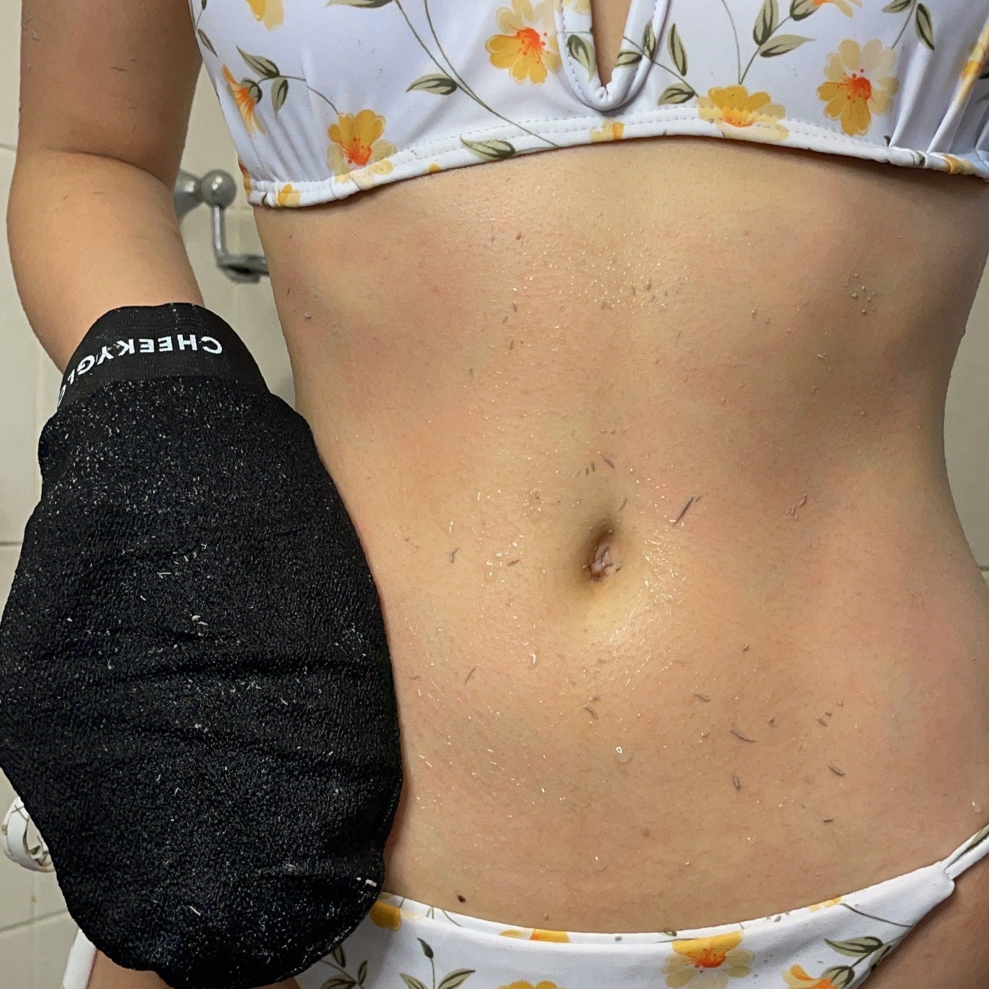 A person, showcasing the CheekyGlo Exfoliating Trio, is displaying a white bikini adorned with yellow and orange flowers. Their torso down to their legs is visible as they hold an exfoliating glove on the left side of their body, revealing small skin flakes. The background features a tiled wall with a visible towel hook.