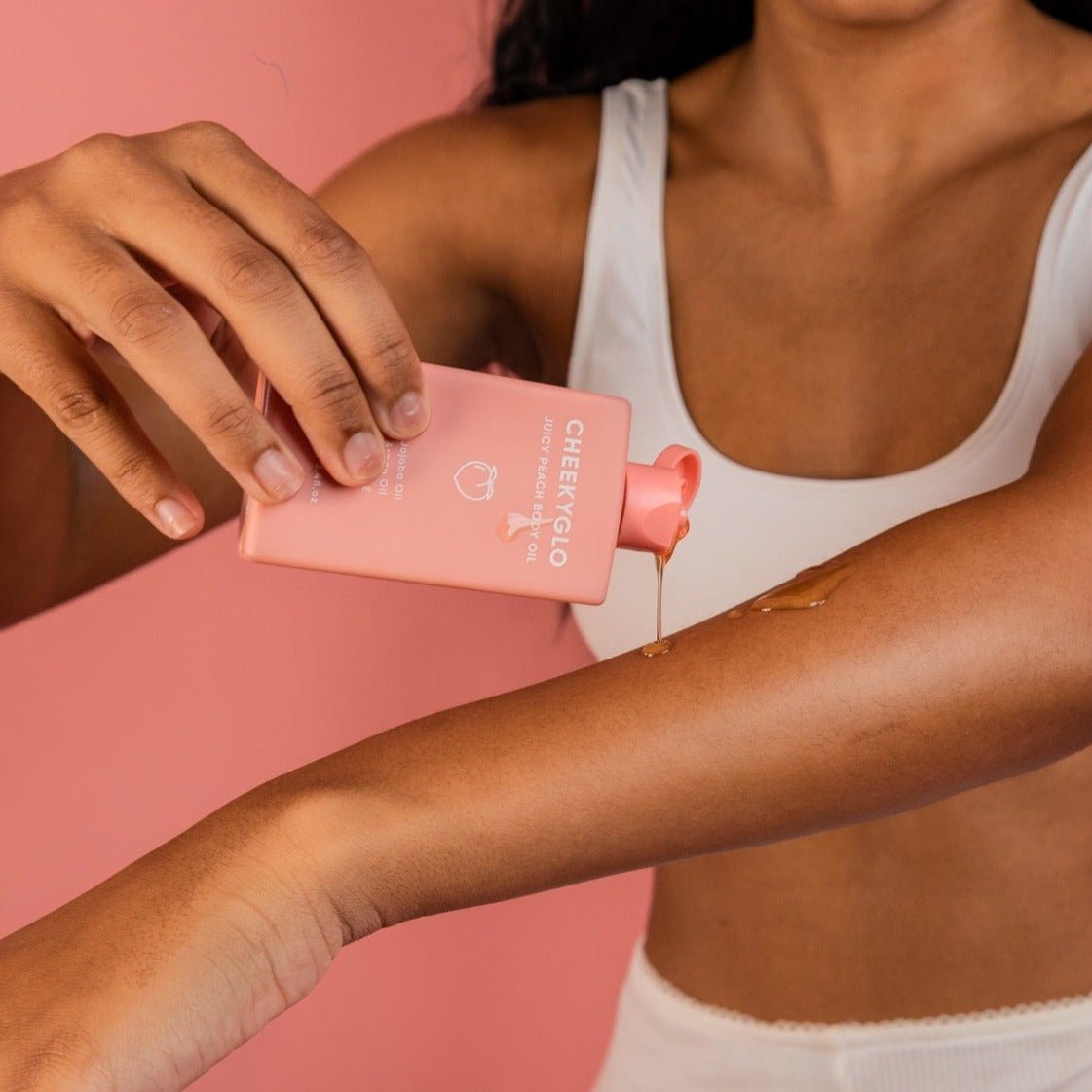 A person with long hair is applying CheekyGlo Juicy Peach Body Oil from a pink tube labeled "CheekyGlo" onto their forearm. They are wearing a white top, and the background is a solid pink color.