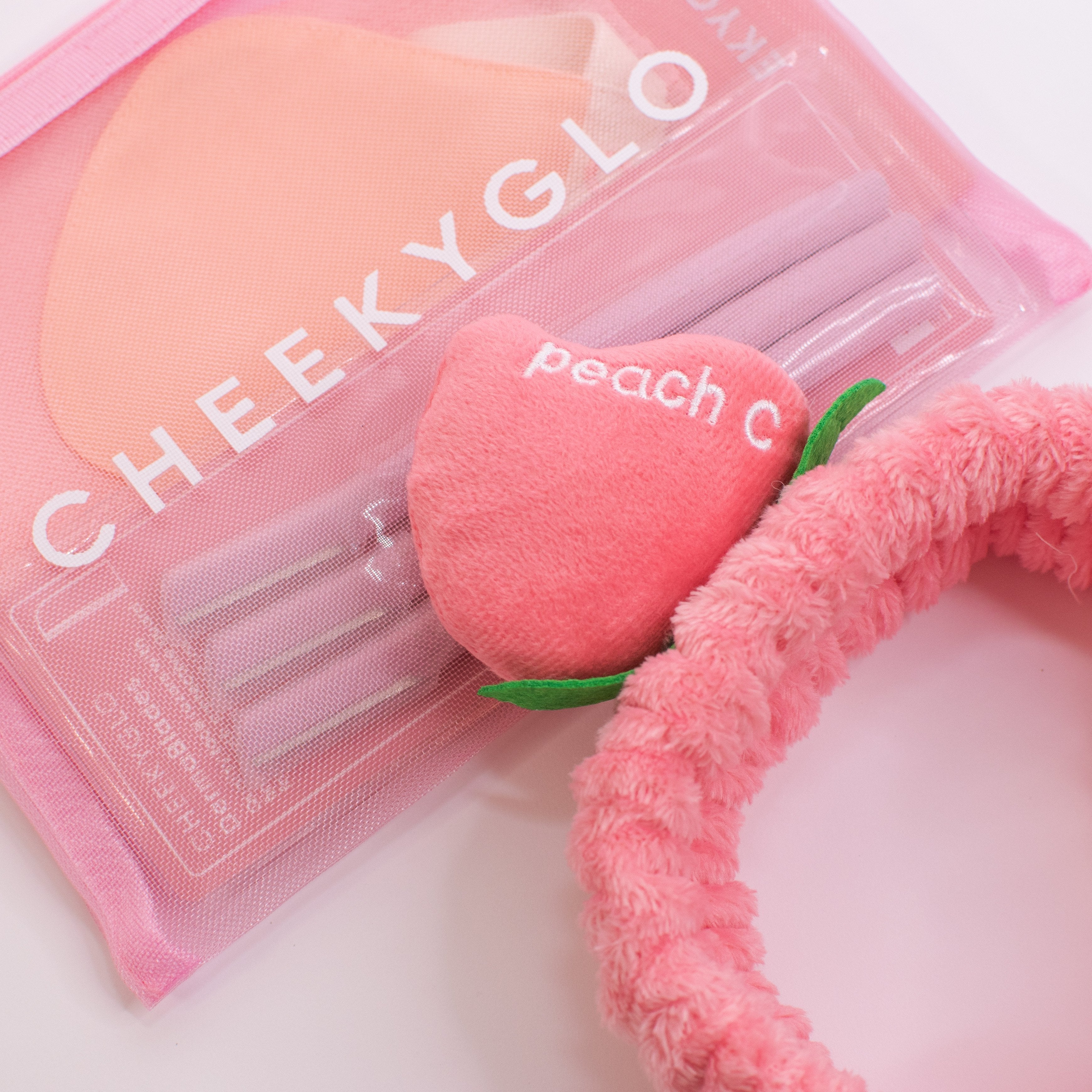 The CheekyGlo Peachy Headband, featuring a plush peach accessory with the word "peach" and the letter "C" embroidered on it, sits next to a pink CHEEKYGLO pouch filled with other items. This fluffy elastic headband is ideal for your self-care routine and looks stunning against a light background.