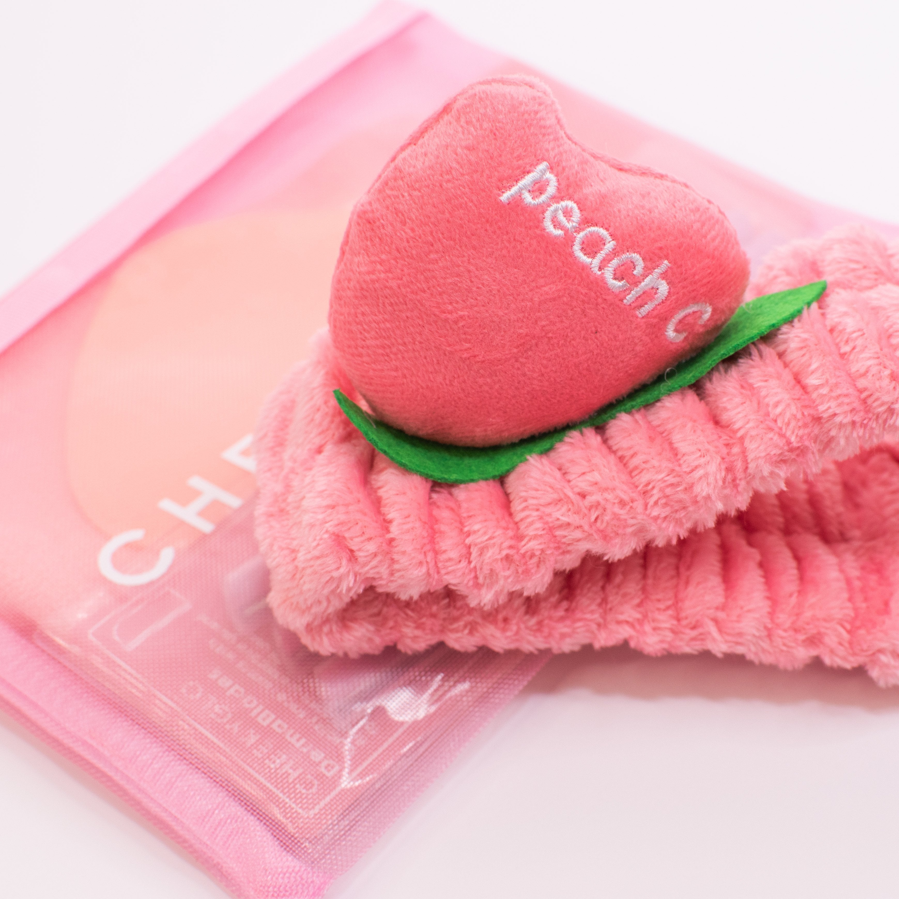 The CheekyGlo Peachy Headband by CheekyGlo is an ultra-soft and plush hairband, featuring a peach-shaped adornment embroidered with "peach c." Both the fluffy elastic headband and its adornment are vibrant pink with green leaves, making it ideal for your self-care routine. The set is elegantly presented on a pastel pink zippered pouch with subtle text that reads "CHE.