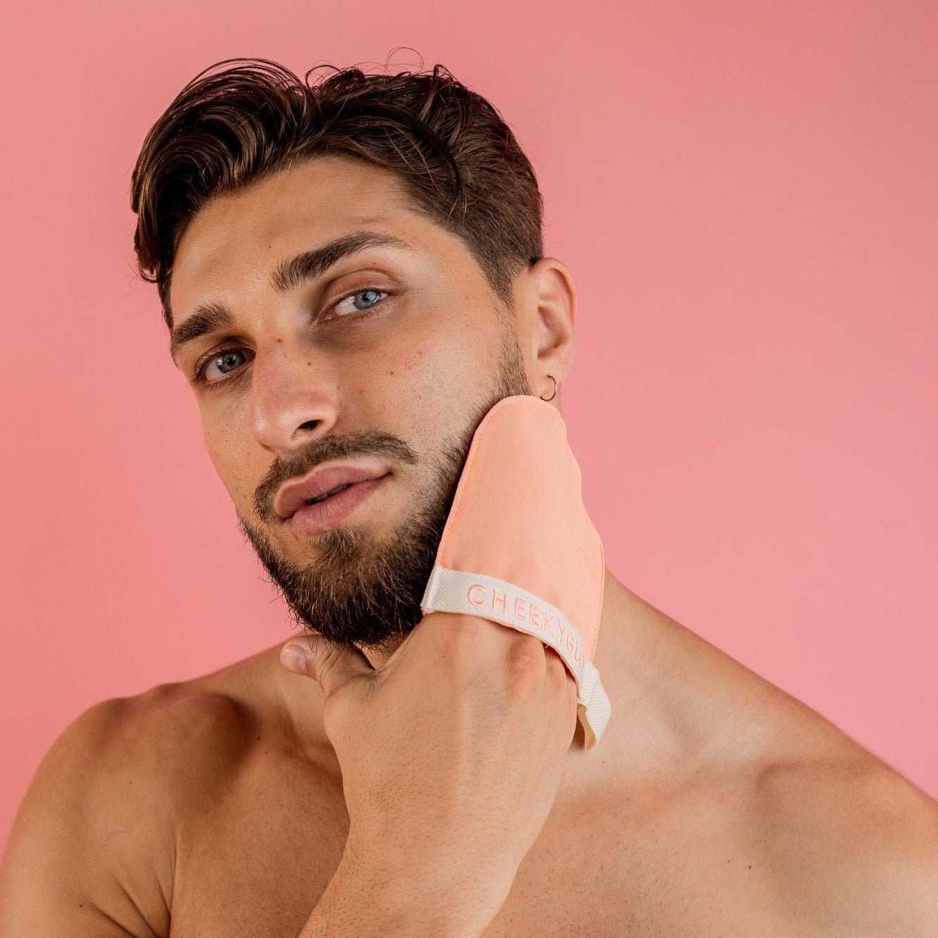 A bearded man with short hair uses a peach-colored CheekyGlo Silk Exfoliating Face Mitt on his cheek against a pink background. The mitt, made from Premium Turkish Silk, has the text "CHEEKY" visible on it. The man&