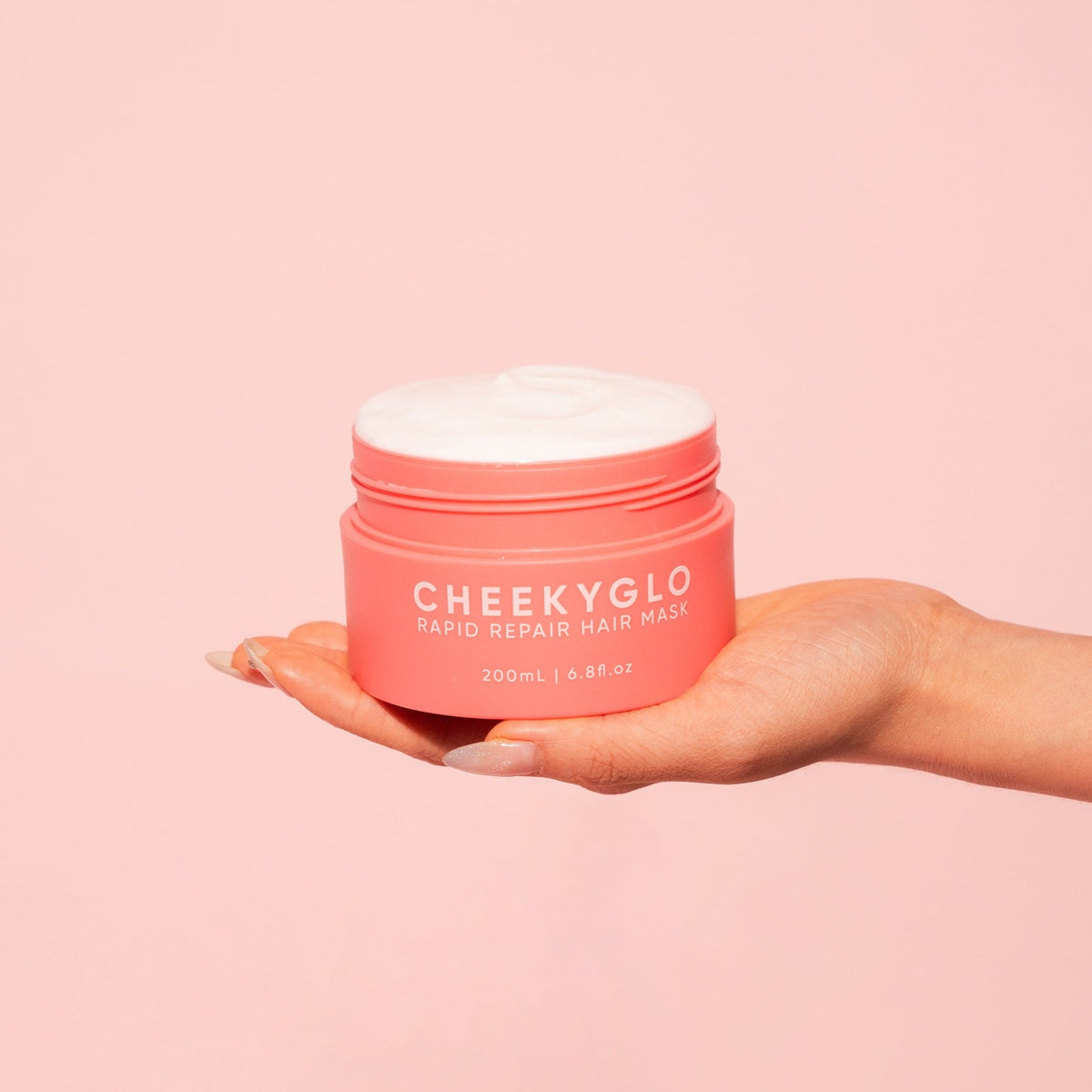 A person holds a container of CheekyGlo's Rapid Repair Hair Mask against a peach-colored background. The pink, slightly opened container reveals the white, creamy hair mask inside. The label indicates that the container holds 200ml (6.8 fl. oz.) of nourishing ingredients designed to leave hair smooth and shiny.