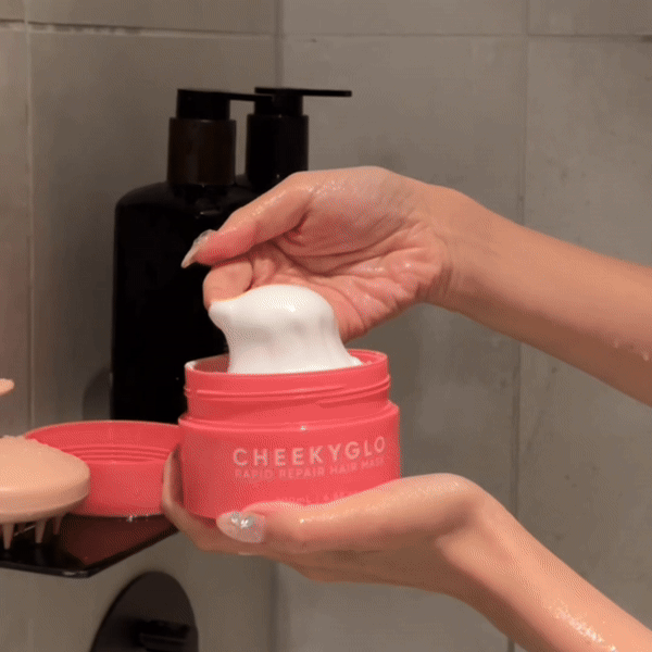 A person scoops a generous amount of white hair mask from a coral-colored container labeled "CheekyGlo Rapid Repair Hair Mask," known for its nourishing ingredients that promote smooth and shiny hair. The container sits next to several black dispenser bottles in a bathroom setting.