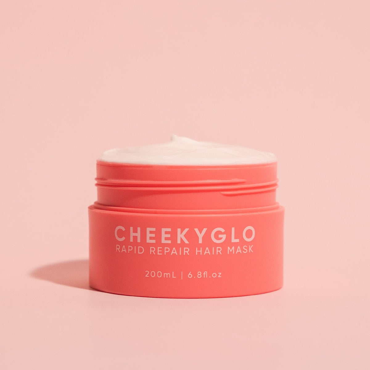 A pink container of CheekyGlo's Rapid Repair Hair Mask is centrally placed against a lighter pink background. The lid is off, revealing a white, creamy substance designed to provide smooth and shiny hair with its nourishing ingredients. The container text states "200mL / 6.8fl.oz.