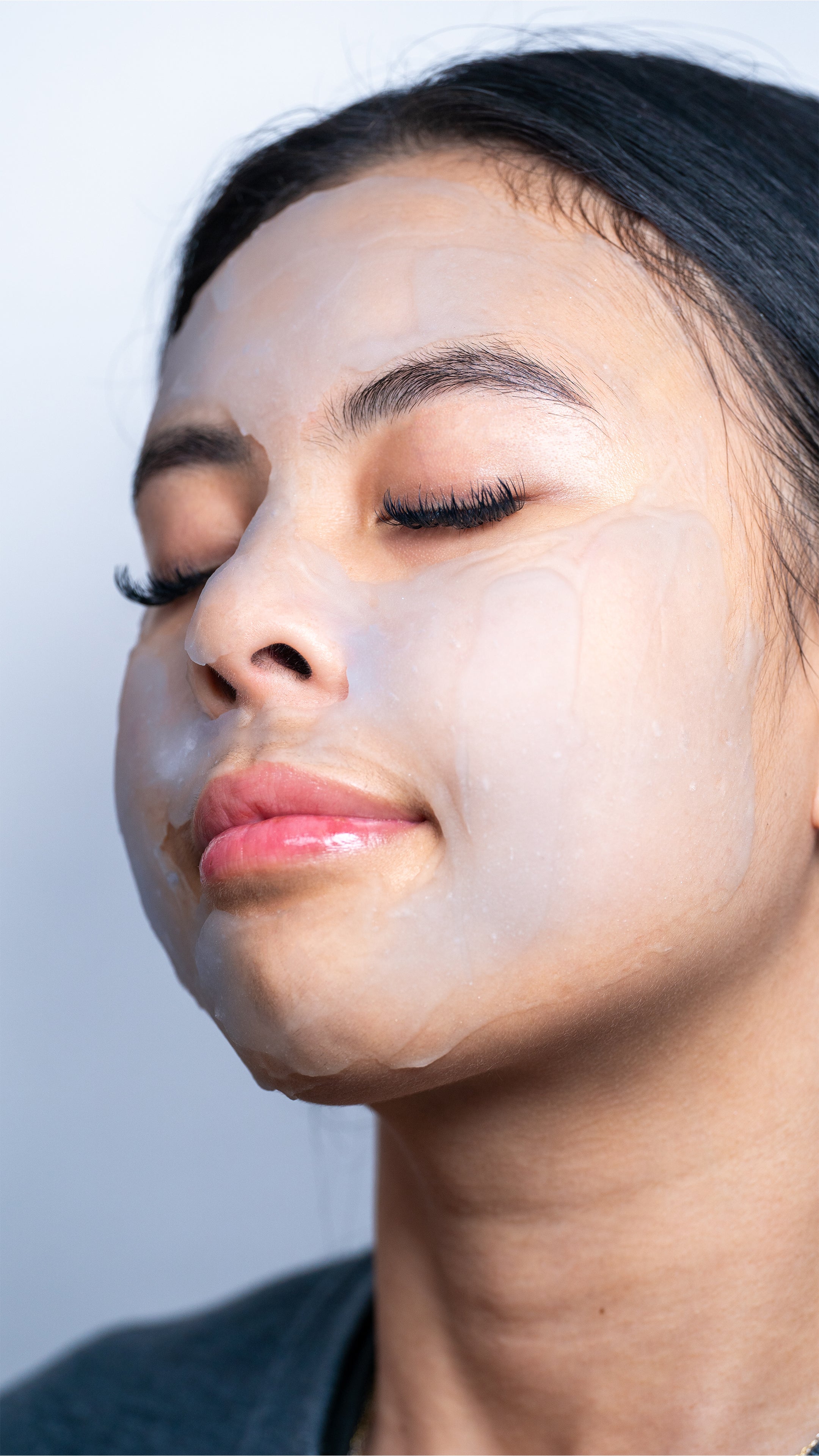 A relaxed person with closed eyes wears the translucent CheekyGlo Ice Slushy Soothing Mask by CHEEKYGLO, suggesting a skincare routine. Their hair is pulled back, and their lips are slightly parted, conveying tranquility and self-care.