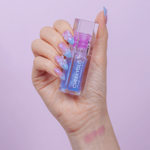 A hand with long, colorful, and glossy pointed nails holds a small clear bottle of CheekyGlo's Colour Changing Lip Oil against a light purple background. Featuring pH-powered technology, the colour-changing lip oil adapts to your natural lips for a personalized hue. The nails boast blue and pink hues with a shiny, intricate design.