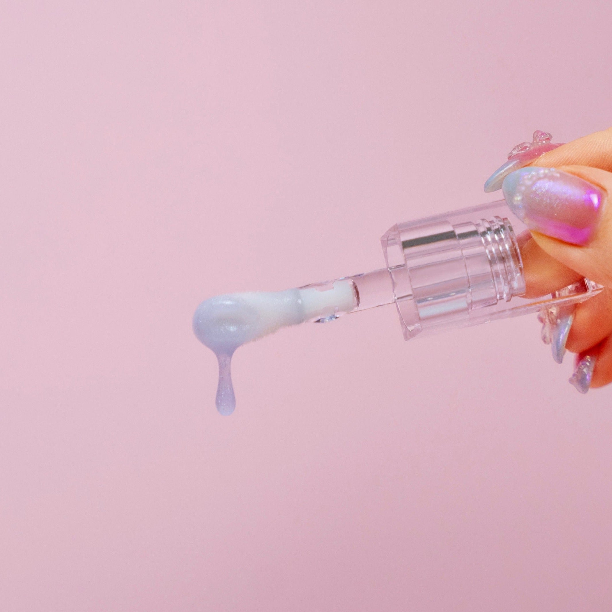 A hand with iridescent nails holds a CheekyGlo Colour Changing Lip Oil (Pink), with a droplet glistening from the applicator. Set against a solid pastel pink background, the scene exudes a soft and colorful aesthetic.