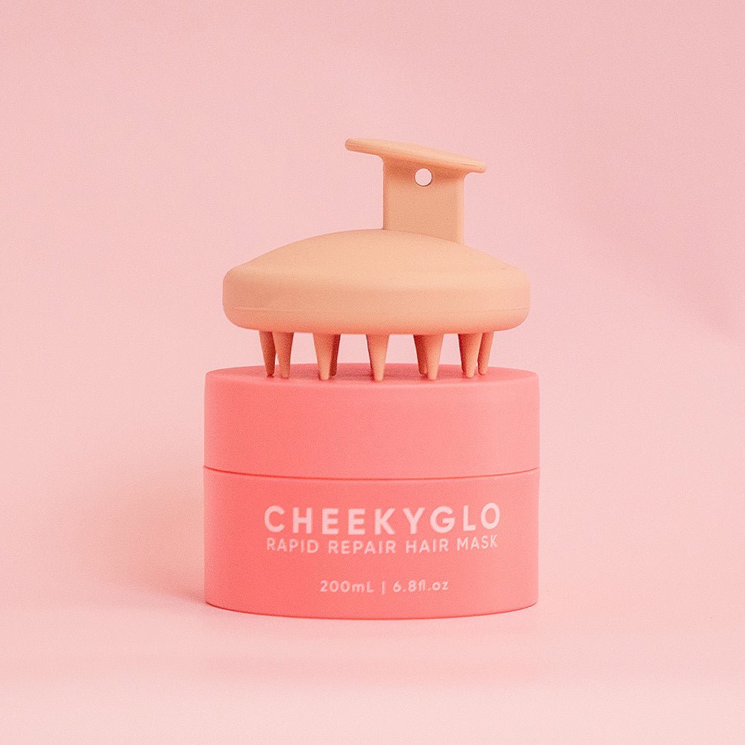 A pink cylindrical container labeled "Hair &amp; Scalp Set" from CheekyGlo, featuring the "RAPID REPAIR HAIR MASK 200mL | 6.8 fl. oz," is topped with a beige scalp massager with short, pointed bristles—an ideal tool for addressing product build-up on hair and scalp. The background is a solid, light pink color.