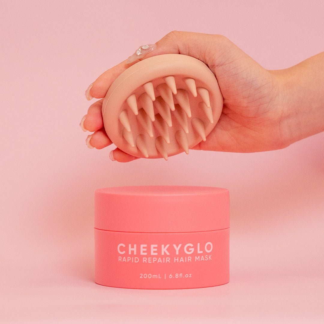 A hand holding a pink scalp massager above a matching pink container of &quot;CheekyGlo Hair &amp; Scalp Set,&quot; suitable for all hair types. The 200ml / 6.8fl.oz container helps remove product build-up, ensuring healthy hair and scalp. Both the background and objects are pink, creating a cohesive color theme.