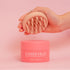 A hand holding a pink scalp massager above a matching pink container of "CheekyGlo Hair & Scalp Set," suitable for all hair types. The 200ml / 6.8fl.oz container helps remove product build-up, ensuring healthy hair and scalp. Both the background and objects are pink, creating a cohesive color theme.