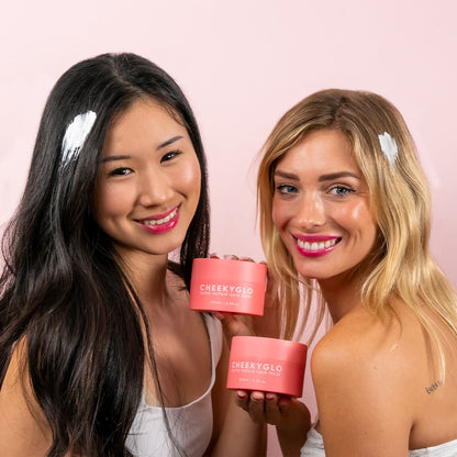 Two smiling women hold peach-colored containers labeled &quot;CheekyGlo,&quot; standing against a light pink background. Each has smooth and shiny hair with a small patch of white cream. The containers are labeled with white text, hinting at the Rapid Repair Hair Mask inside, enriched with nourishing ingredients.