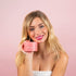 A smiling woman with long blonde hair holds up a pink product box labeled "Rapid Repair Hair Mask" by CheekyGlo against a matching pink background. She has minimal makeup, pink lipstick, and is wearing a white strapless top. Her smooth and shiny hair frames an intricate tattoo visible on her left shoulder.