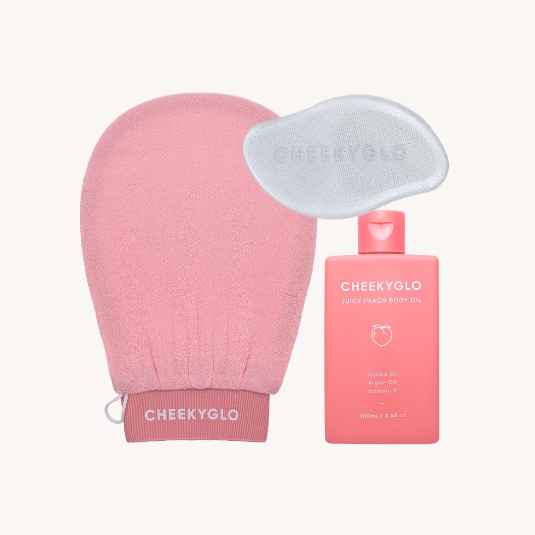 A whimsical CheekyGlo Scrub &amp; Buff Set showcases a pink exfoliating glove, a light blue facial exfoliating pad, and a foot file next to a pink bottle of CheekyGlo Juicy Peach Body Oil. The body oil bottle is adorned with a minimalist peach illustration and text highlighting key ingredients like Jojoba Oil, Argan Oil, and Vitamin E.