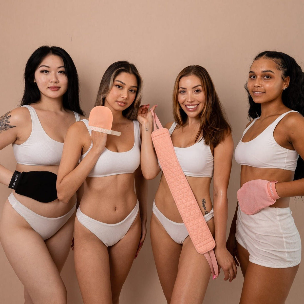 Four women in matching white outfits hold CheekyGlo's Ultimate Glow Set + Free Makeup Bag, featuring exfoliating gloves and body scrubbers. They stand against a beige backdrop, smiling at the camera and showcasing skincare products designed for glowing skin.