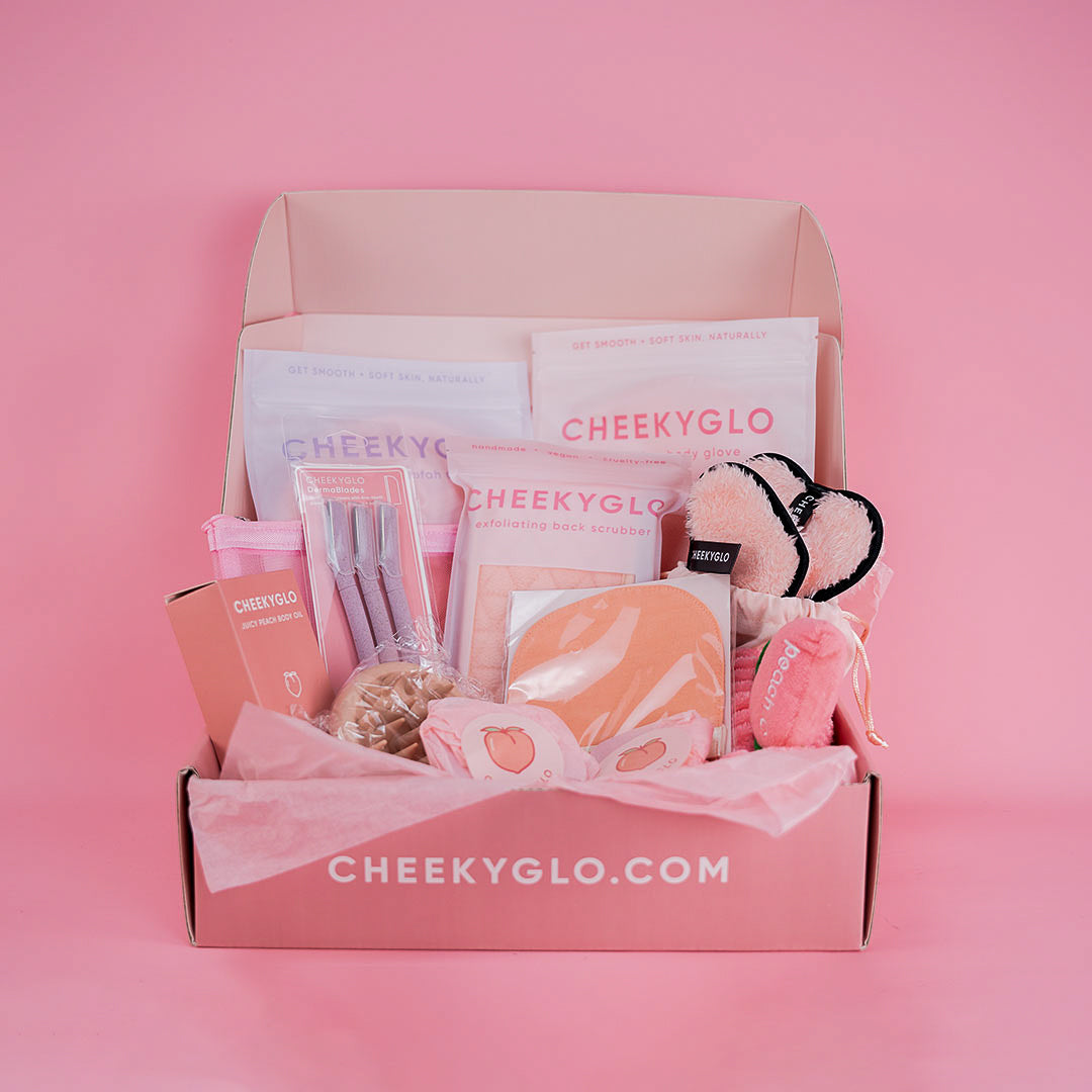 A pink open box with &quot;cheekyglo.com&quot; on the flap contains The Everything Set [BEST VALUE] by CheekyGlo. Visible items include an exfoliating body glove, skincare packets, a hairband, and other beauty accessories, all arranged neatly on pink tissue paper. The background is a complementary shade of pink.