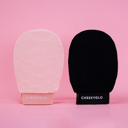 The Black+Pink Exfoliating Glove Duo by CheekyGlo is showcased against a pink background. The set includes one light pink mitt and one black mitt, both featuring straps at the base with &quot;CheekyGlo&quot; printed in white, promising smoother skin with every use.