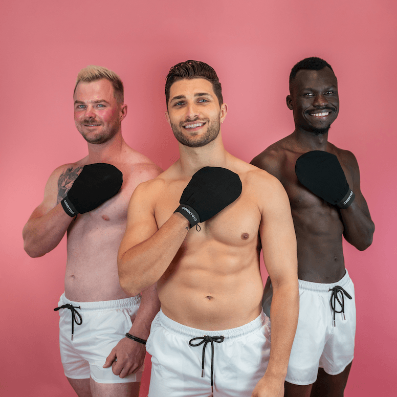 Three smiling men, all shirtless and wearing white shorts, stand against a pink background. Each holds a black exfoliating glove from the CheekyBro Set by CheekyGlo to their chest. The men&