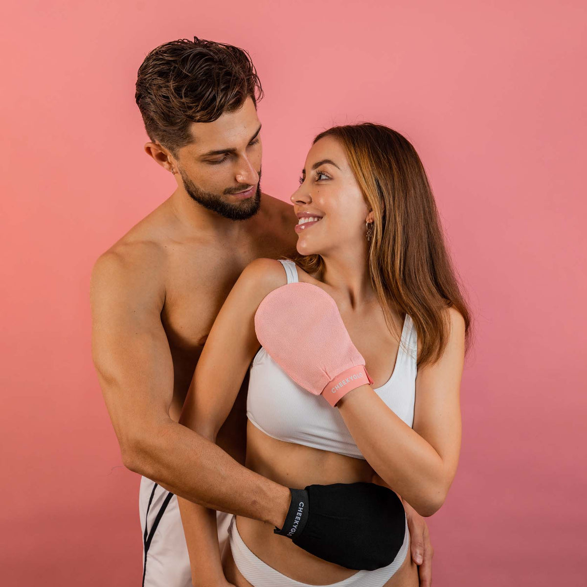 A shirtless man stands behind a woman wearing white underwear and a tank top. Both are showcasing the Black+Pink Exfoliating Glove Duo by CheekyGlo, with the woman's glove held to her chest and the man's glove resting on his waist. They share a smiling, affectionate gaze against a pink background, ready to achieve smoother skin together through effective dead skin removal.