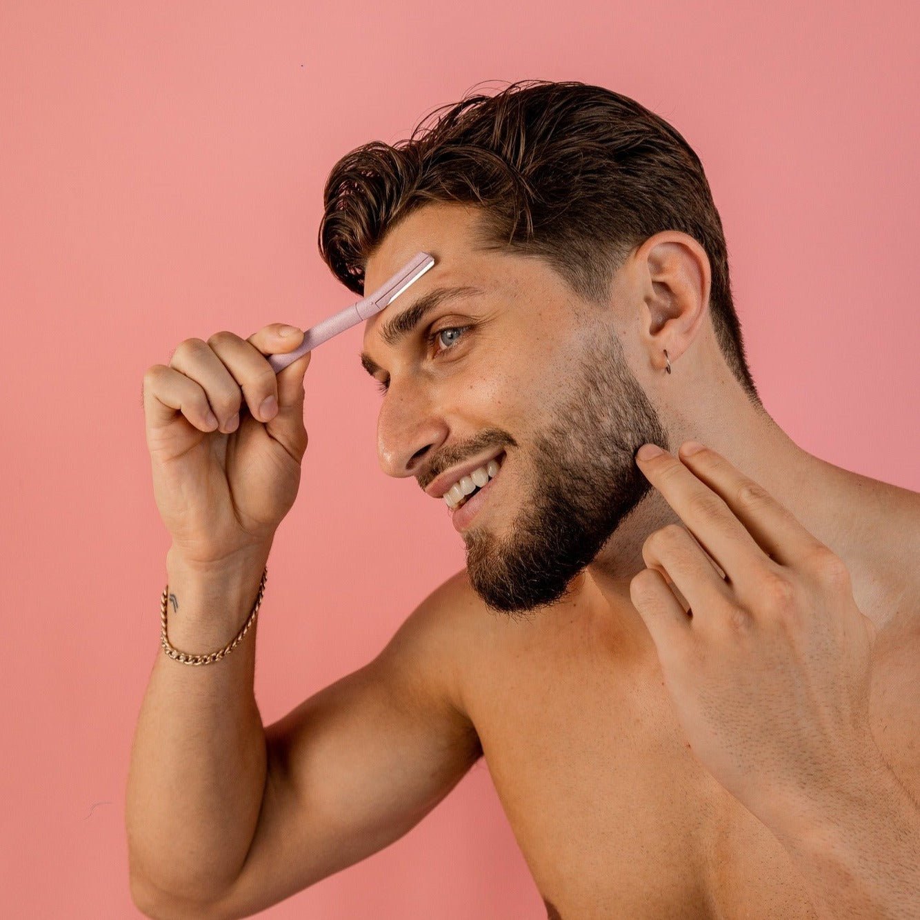 A smiling, shirtless man with dark hair and a beard stands against a pink background, using CheekyGlo Dermablades from CheekyGlo to groom his eyebrows. Wearing a gold bracelet and stud earring, he holds the skin on his left eyebrow taut with his fingers while using the eco-friendly razor.