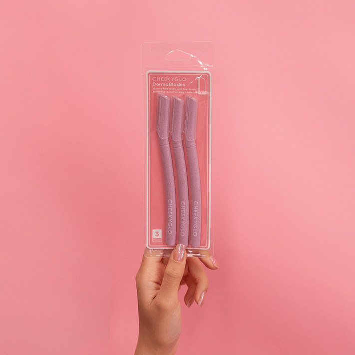 A hand holds a clear plastic package containing three CheekyGlo Dermablades, designed for facial hair removal, against a solid pink background. The package showcases the brand name &quot;CheekyGlo&quot; along with product details, highlighting their eco-friendly blades.