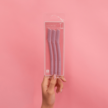 A hand holds a clear plastic package containing three CheekyGlo Dermablades, designed for facial hair removal, against a solid pink background. The package showcases the brand name &quot;CheekyGlo&quot; along with product details, highlighting their eco-friendly blades.