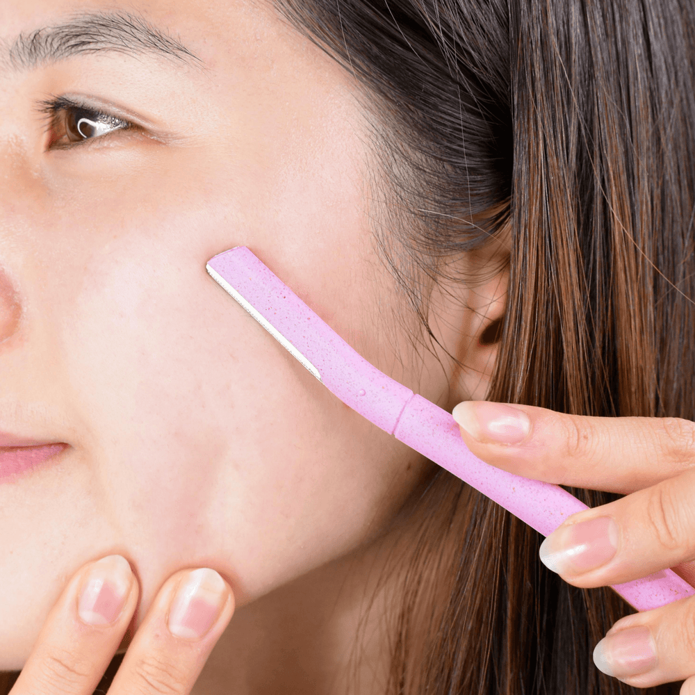 Close-up of a person using a CheekyGlo Dermablade for facial hair removal from their cheek. The person's skin looks smooth as they gently pull the skin taut with their fingers while using the eco-friendly blade from CheekyGlo.