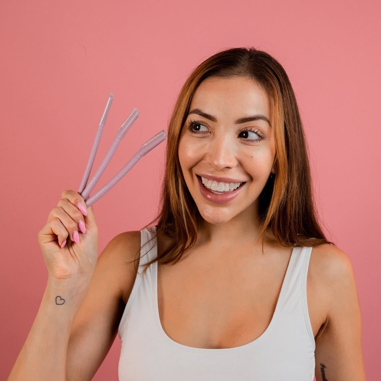 A smiling woman with long brown hair holds three CheekyGlo Dermablades in her right hand against a pink background. She is wearing a white tank top and has a small heart tattoo on her left wrist.