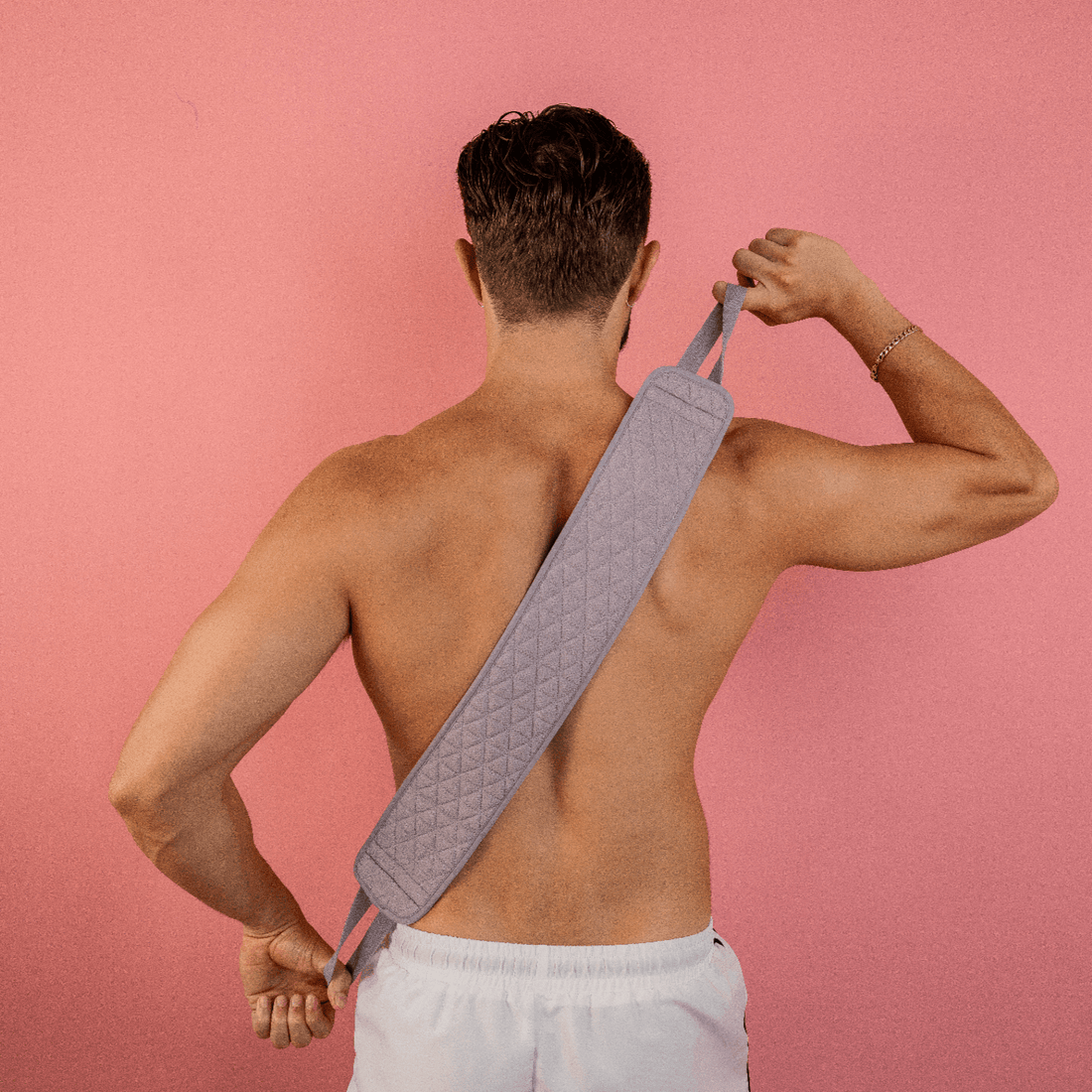 A shirtless man stands in front of a pink background, wearing white shorts while using the CheekyGlo Exfoliating Back Scrubber to massage his congested skin. Made from biodegradable viscose, he grips the gray textured strap with both hands.