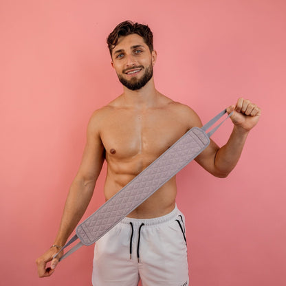 A shirtless man with a beard holds a quilted gray exercise band against a pink background, featuring his CheekyGlo Exfoliating Back Scrubber. He wears white shorts, smiles at the camera, and enjoys the fresh feel of biodegradable viscose from CheekyGlo to relieve congested skin.