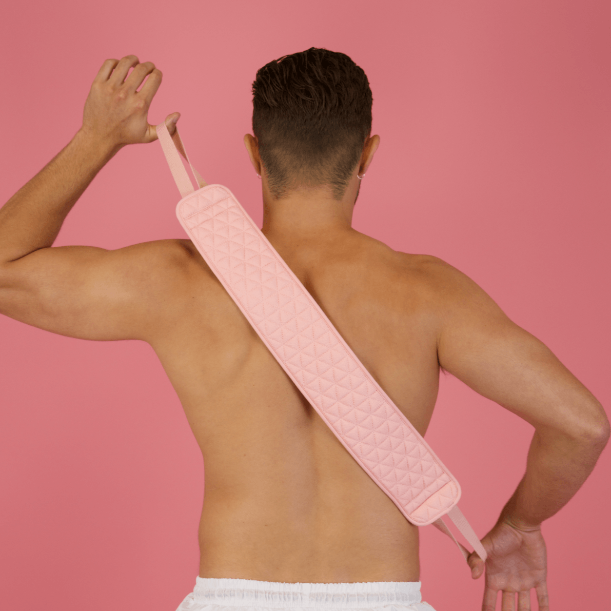 A person with short hair, viewed from behind, uses a CheekyGlo Exfoliating Back Scrubber crafted from biodegradable viscose, draping it diagonally across their back. They are positioned against a pink background and dressed in white shorts.