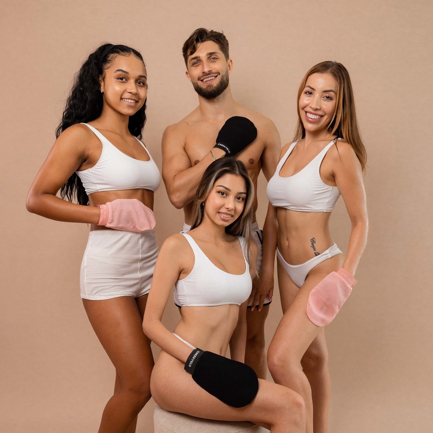 Four people, three women and one man, pose against a beige background wearing white sportswear. They are holding CheekyGlo Exfoliating Gloves in different colors—pink and black—and smiling confidently. The group appears healthy and fit, ready for smoother, healthier skin with their CheekyGlo products.