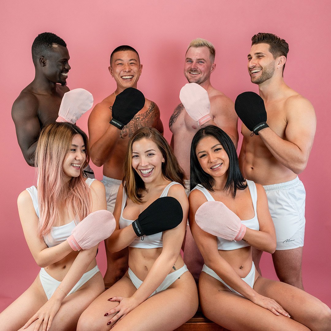 A diverse group of three men and three women pose cheerfully against a pink background, wearing minimal white attire and holding CheekyGlo Exfoliating Gloves. The men display black mitts while the women have pink ones. Smiling brightly, they showcase their journey towards smoother, healthier skin with CheekyGlo.