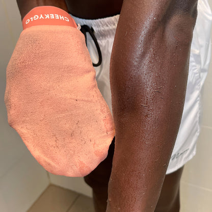 A person wearing white shorts is using a CheekyGlo Exfoliating Glove on their right arm. The light orange glove, branded &quot;CheekyGlo&quot; on the wristband, glides over damp skin. The bright lighting accentuates the textures on the arm and glove, promising smoother and healthier skin with each use.