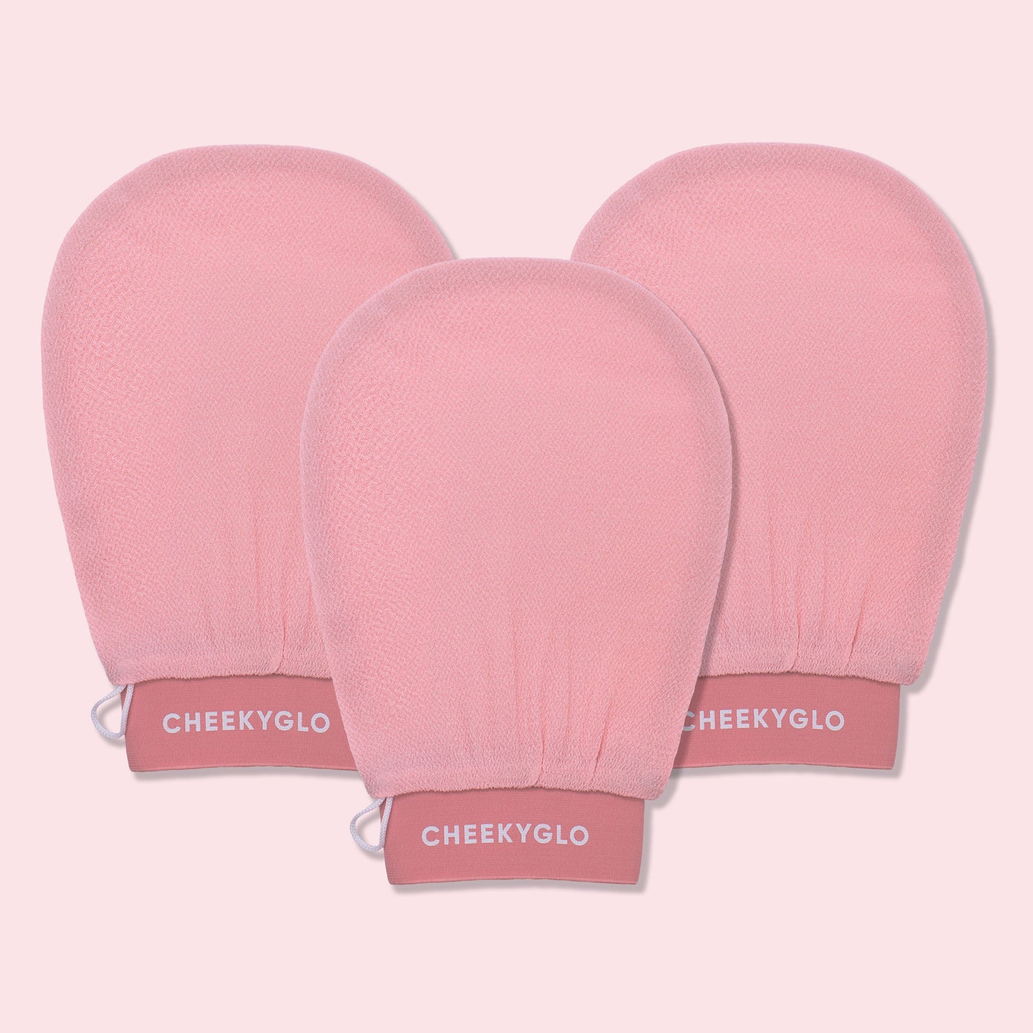 Three CheekyGlo Exfoliating Gloves are displayed against a light pink backdrop. The gloves are slightly rounded at the top with elasticated wristbands labeled &quot;CHEEKYGLO,&quot; promising smoother skin and a healthier glow.