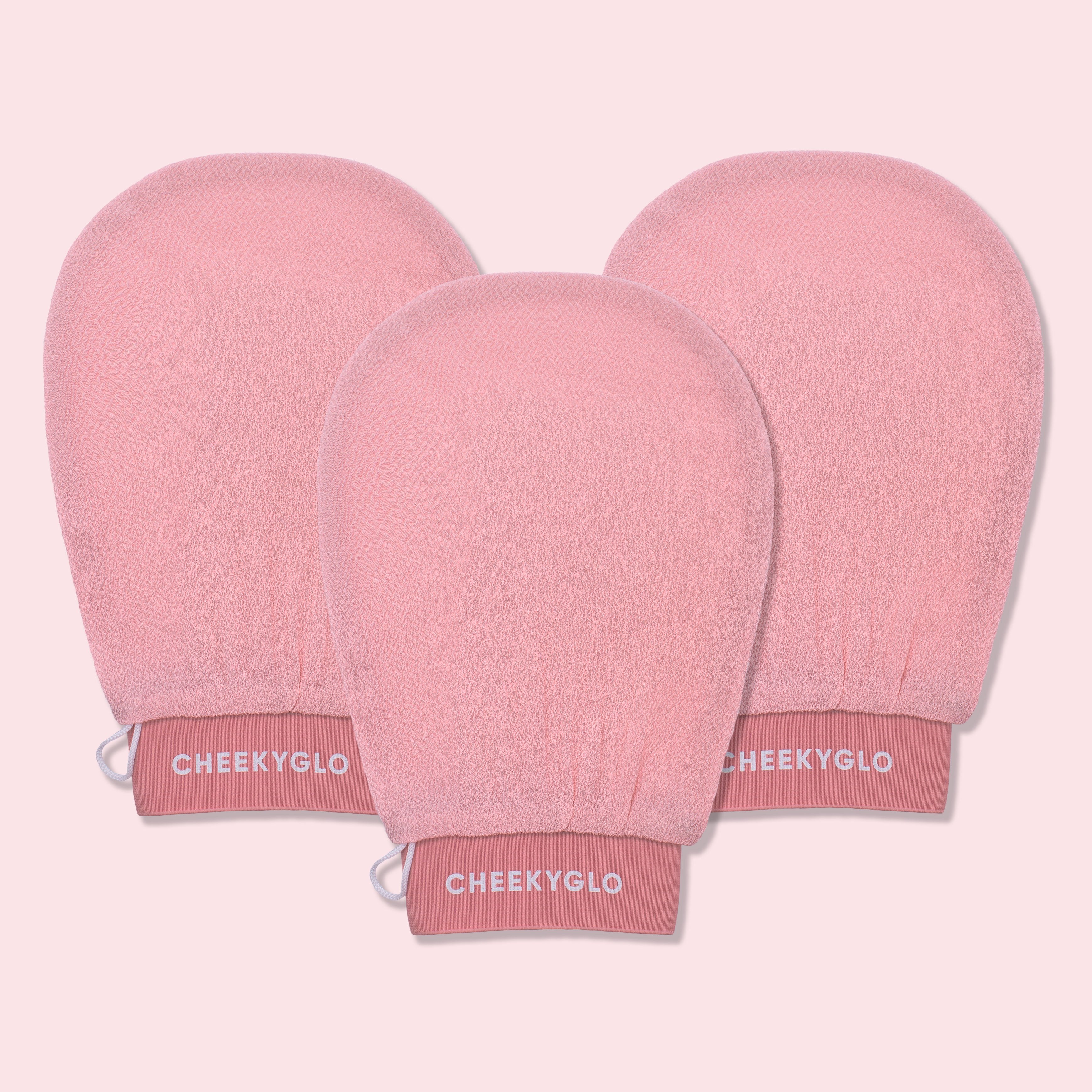 Three CheekyGlo Exfoliating Gloves are displayed against a light pink backdrop. The gloves are slightly rounded at the top with elasticated wristbands labeled &quot;CHEEKYGLO,&quot; promising smoother skin and a healthier glow.