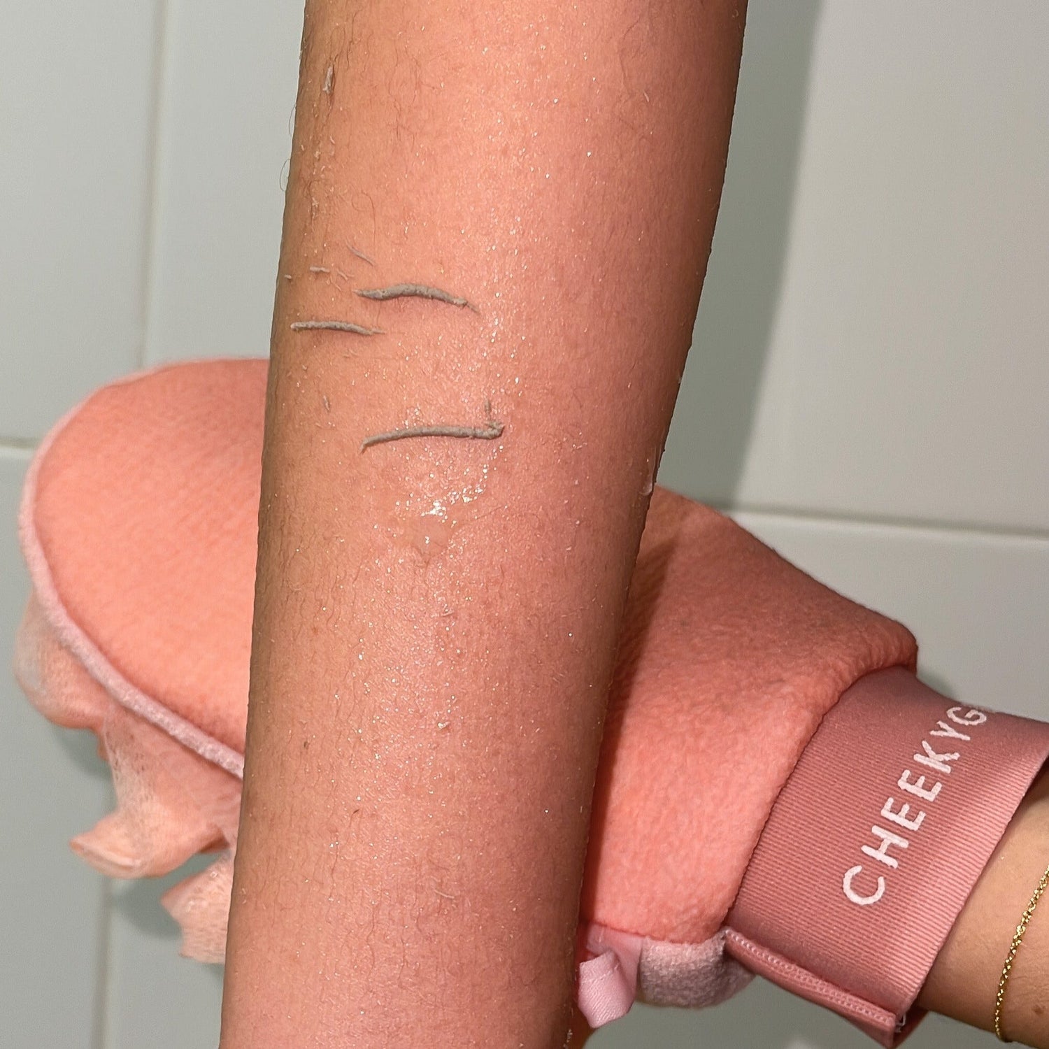 A person holding up their arm, freshly exfoliated with a pink CheekyGlo Exfoliating Loofah Glove. The wet arm displays three small healing cuts on the forearm, indicating effective skin smoothing. A tiled wall in the background suggests they might be in a bathroom, potentially treating keratosis pilaris.