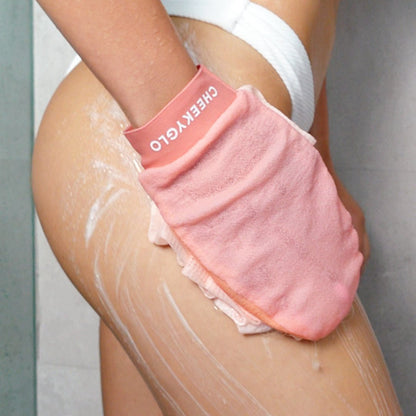 A person is using a pink CheekyGlo Exfoliating Loofah Glove to scrub their upper thigh and hip area while in the shower, aiming for skin smoothing and reducing keratosis pilaris. They are wearing white underwear, with soap or body wash lathered on their skin.