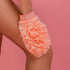 A person using a soft, peach-colored CheekyGlo Exfoliating Loofah Glove with a ruffled texture on their leg. The glove, branded "CheekyGlo" on the wrist strap, promises to help with keratosis pilaris and leave the skin smooth. The background is a solid pink color.