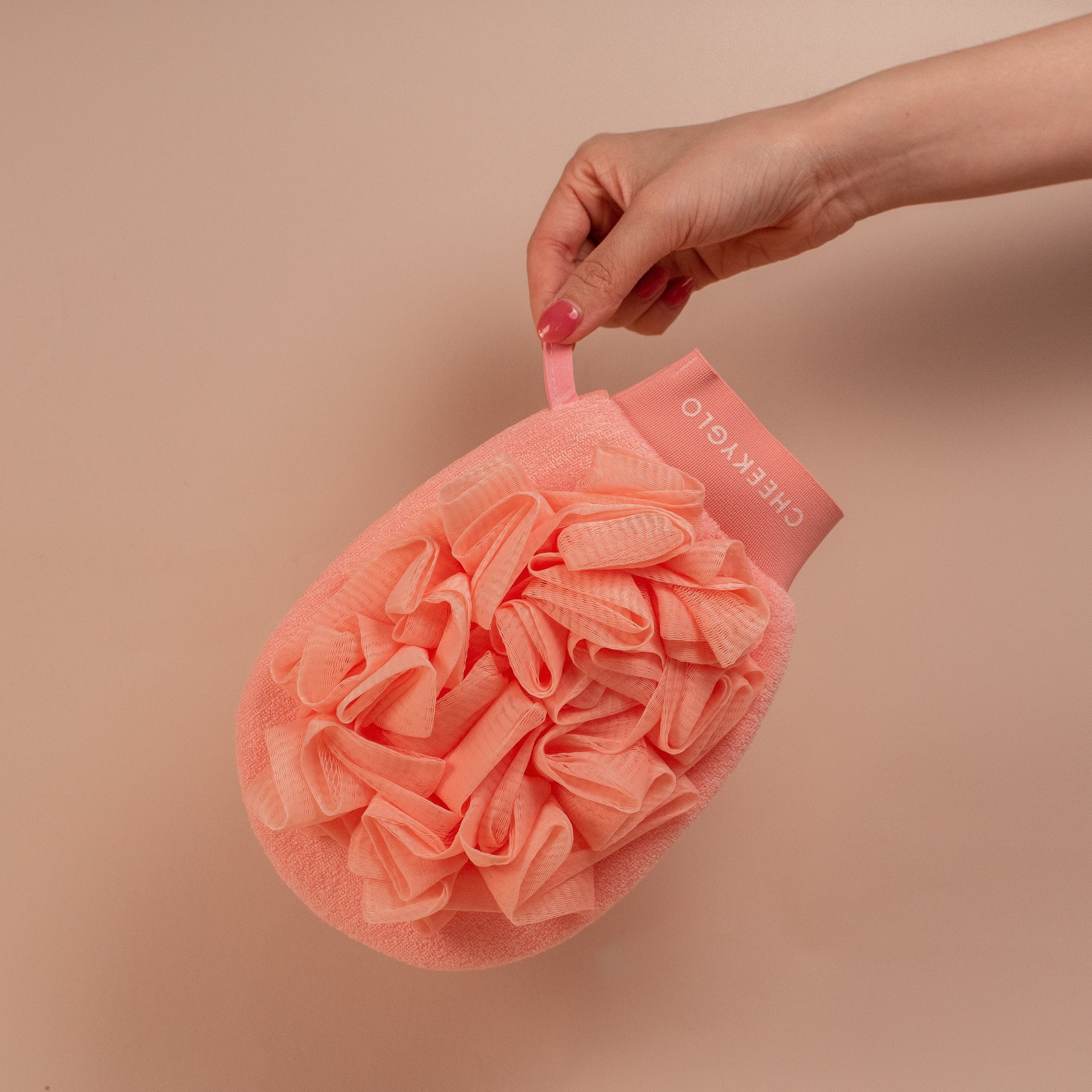 A hand holding a pink CheekyGlo Exfoliating Loofah Glove with a strap labeled "CHEEKYGLO." The glove features a textured, ruffled surface and an oval shape. Ideal for tackling keratosis pilaris, it promises skin-smoothing results. The background is a plain beige color.