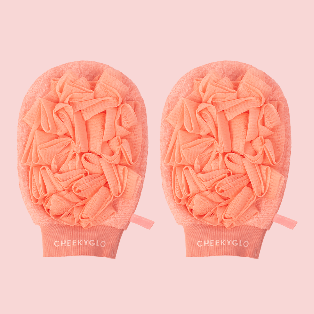 Two coral-pink CheekyGlo Exfoliating Loofah Gloves with ruffled texturing are shown against a matching light pink background. The skin-smoothing gloves feature the brand name &quot;CheekyGlo&quot; near the wrist opening, making them perfect for tackling keratosis pilaris.