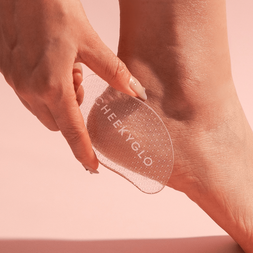 A hand uses the transparent glass foot file &quot;01 Mark - Test&quot; by CheekyGlo for removing dead skin from the heel, set against a soft pink backdrop. It ensures smooth, soft feet with every use.