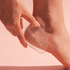 A hand uses the transparent glass foot file "01 Mark - Test" by CheekyGlo for removing dead skin from the heel, set against a soft pink backdrop. It ensures smooth, soft feet with every use.