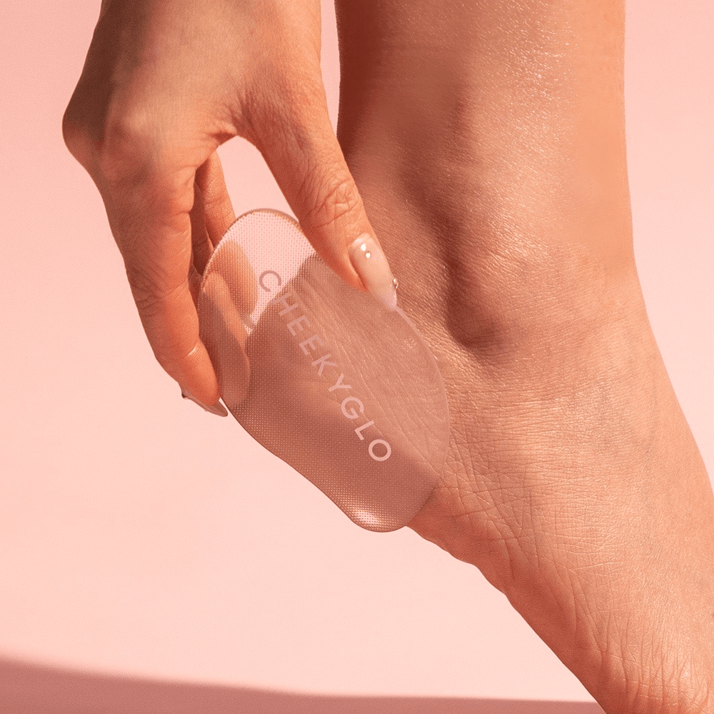 Against a soft pink background, a person uses the "01 Mark - Test" by CheekyGlo, a clear silicone exfoliating pad, on their heel. Their smooth, moisturized skin and gleaming manicured nails highlight its effectiveness in removing dead skin.