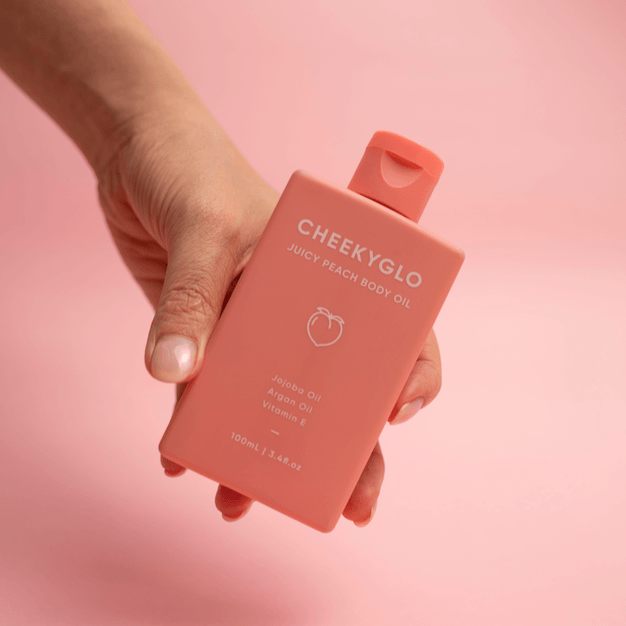 A hand is holding a rectangular, peach-colored bottle labeled &quot;CheekyGlo Juicy Peach Body Oil.&quot; This all-natural body oil from CheekyGlo lists ingredients including Jojoba Oil, Argan Oil, and Vitamin E. The background is a matching peach color.